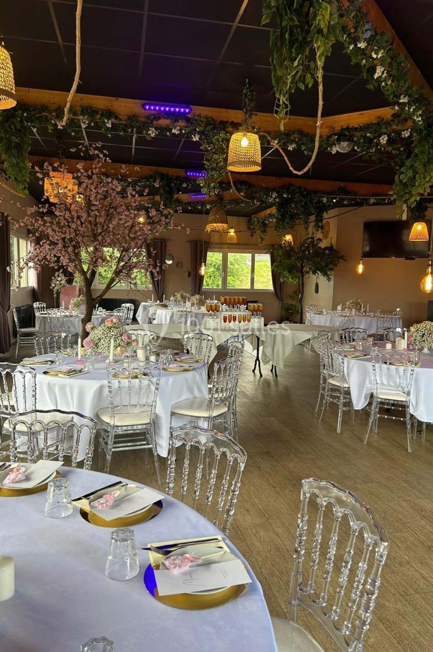 Salle de réception décorée avec des tables rondes dressées, des chaises transparentes et des éléments végétaux suspendus.
