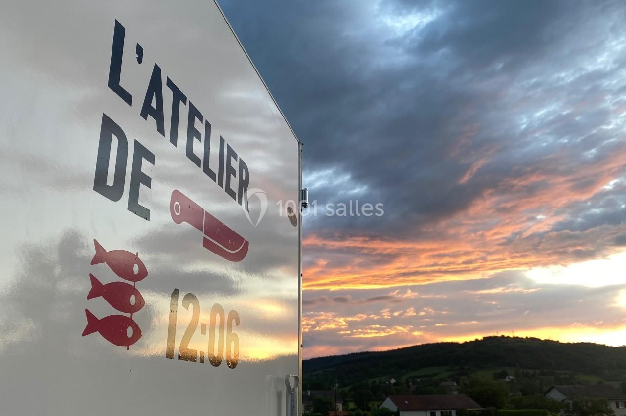 Flanc d'un camion marqué ’L'Atelier de 12:06’ avec un couteau et des poissons, devant un coucher de soleil champêtre.