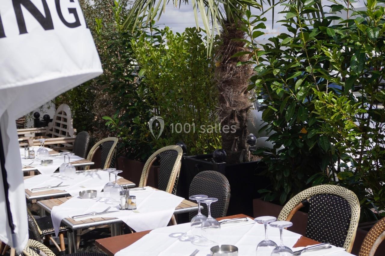 Terrasse de restaurant avec tables dressées, nappes blanches, chaises en osier et végétation en arrière-plan.