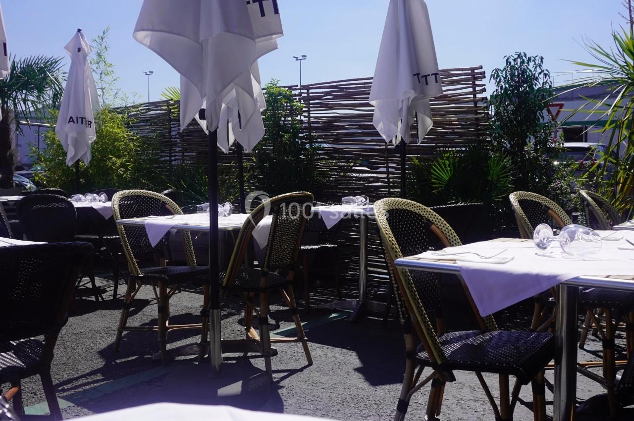 Terrasse ensoleillée d'un restaurant avec tables dressées, chaises en osier et parasols blancs.