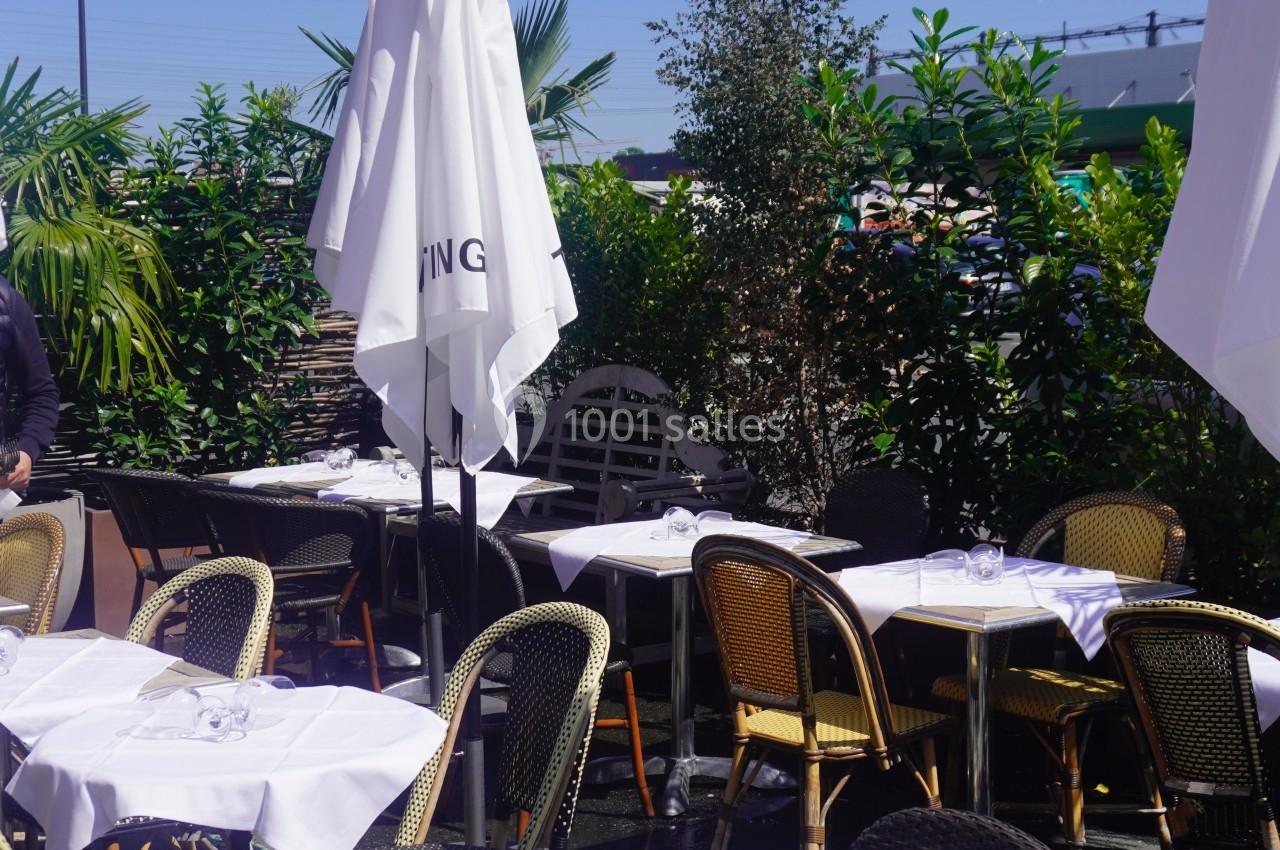 Terrasse ensoleillée avec tables et chaises en osier, nappes blanches et parasols fermés entourés de végétation.