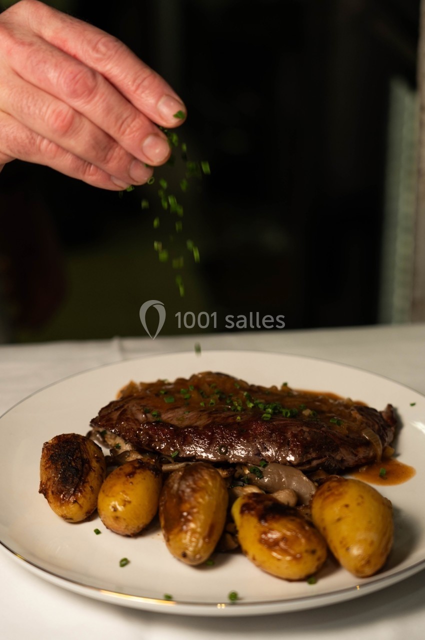 Une main saupoudre de la ciboulette sur un plat de viande en sauce accompagné de pommes de terre rôties.
