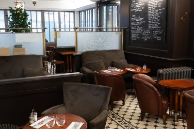 Salle de restaurant avec tables en bois, chaises assorties et grandes fenêtres laissant entrer la lumière naturelle.