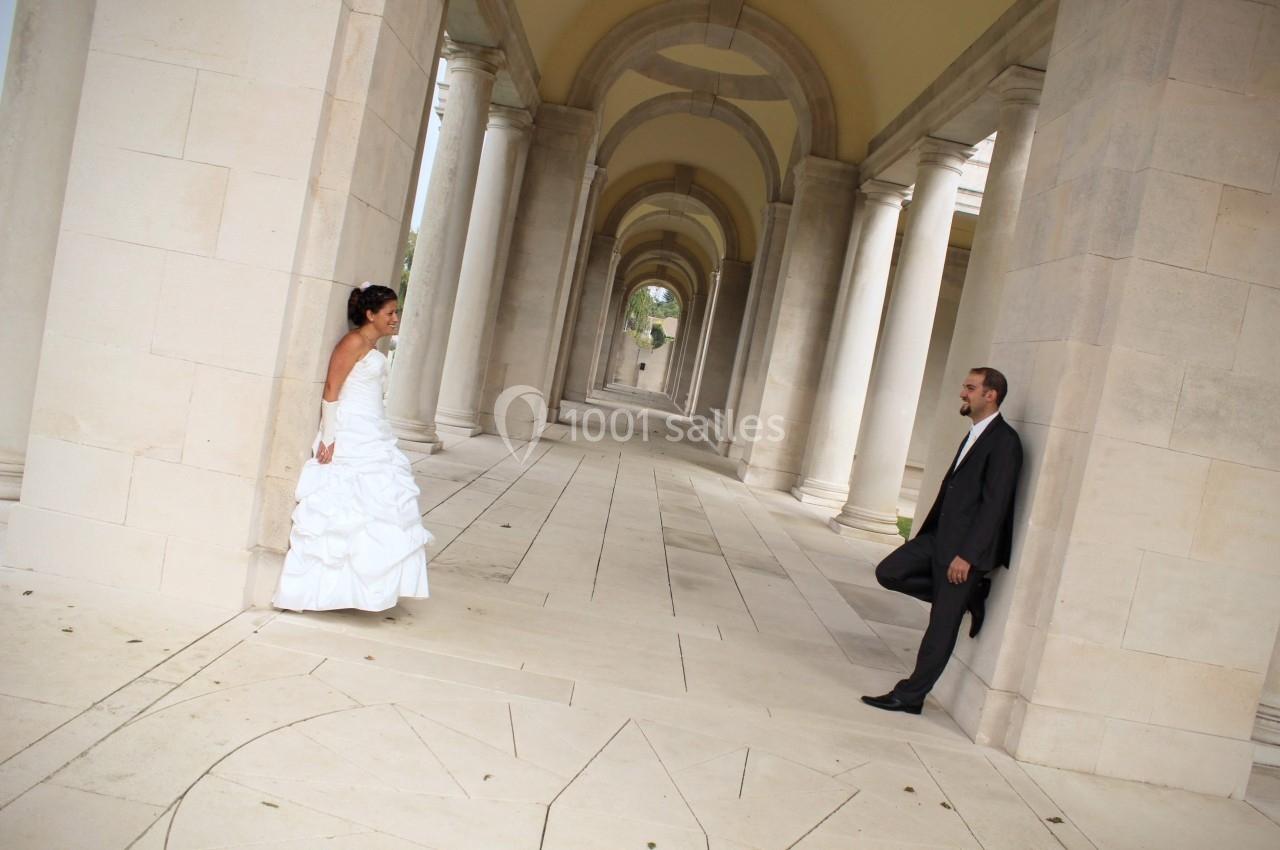 Un couple en tenue de mariage se regarde dans une galerie avec des colonnes et des arches en pierre.