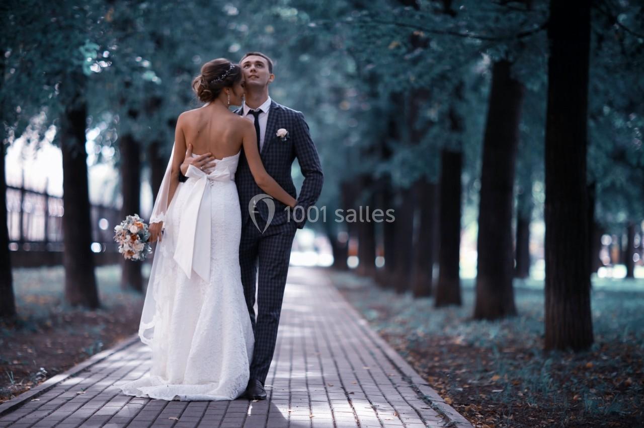 Un couple en tenue de mariage marche sur une allée pavée bordée d'arbres dans un parc.