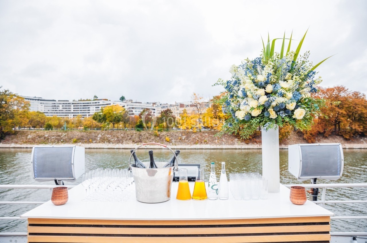 Table avec seau à champagne, verres, carafes de jus et grand bouquet floral, située en extérieur près d'une rivière.