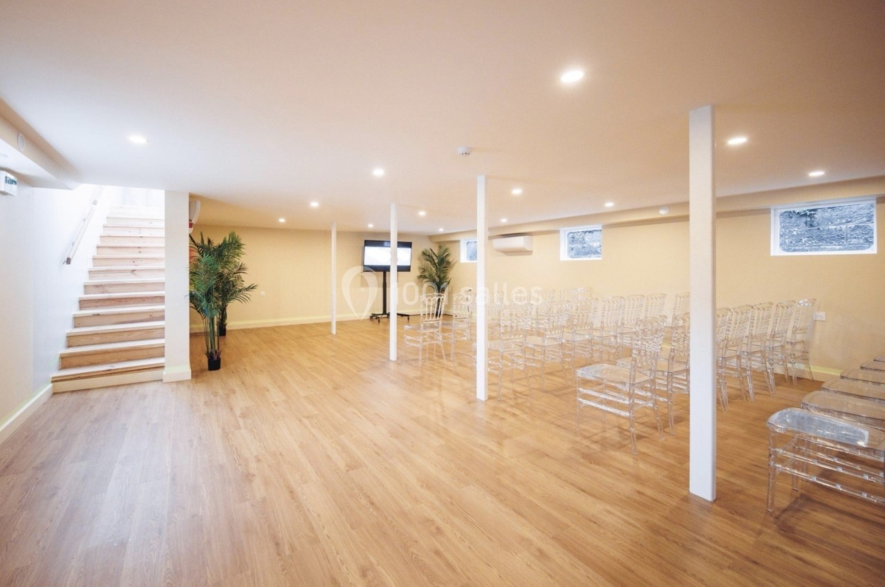 Salle lumineuse avec parquet, chaises transparentes alignées, écran et plantes, escalier menant à l'étage.