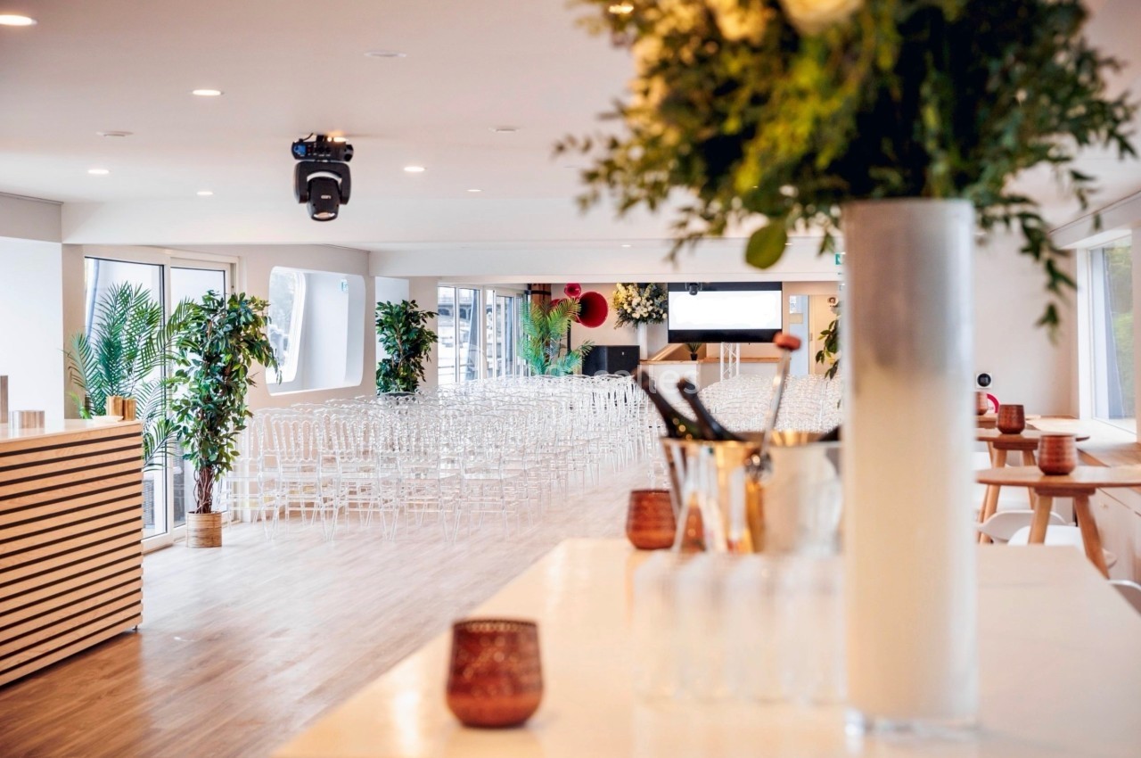 Salle lumineuse aménagée pour un événement, avec des chaises transparentes alignées et des plantes décoratives.