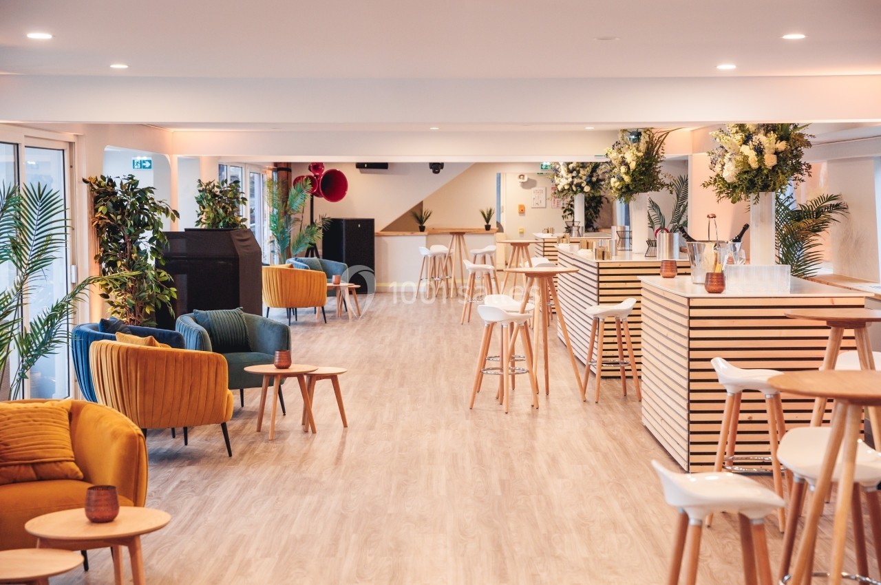 Salle lumineuse avec mobilier moderne, chaises colorées, tables en bois, plantes et comptoirs en bois clair.
