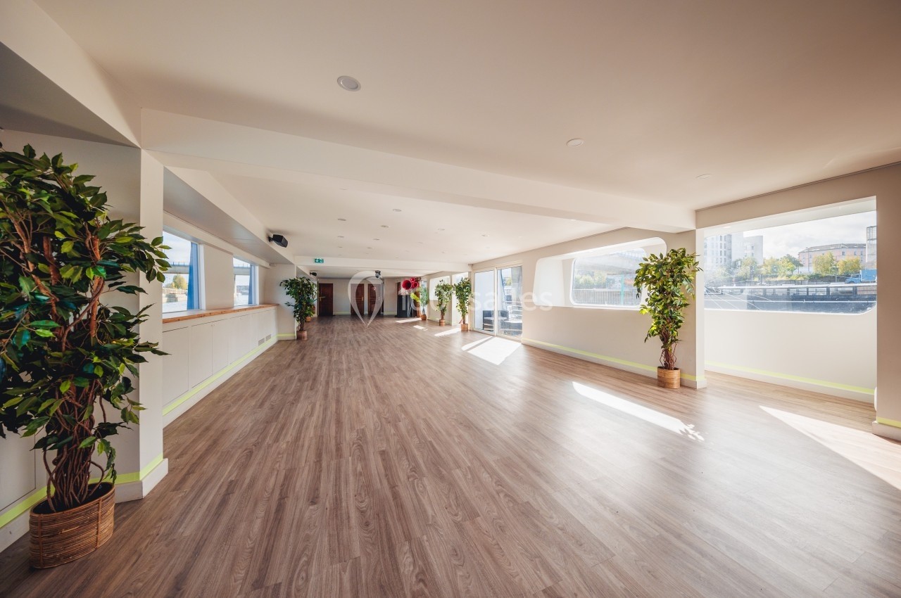 Salle lumineuse avec parquet, grandes fenêtres, plantes en pot et vue sur un extérieur urbain.
