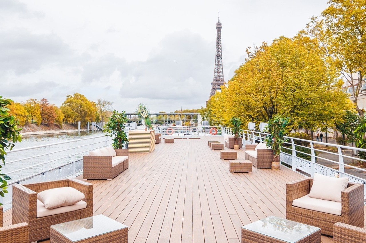 Terrasse en bois aménagée avec des fauteuils et des plantes, offrant une vue sur la Seine et la tour Eiffel en arrière-plan.
