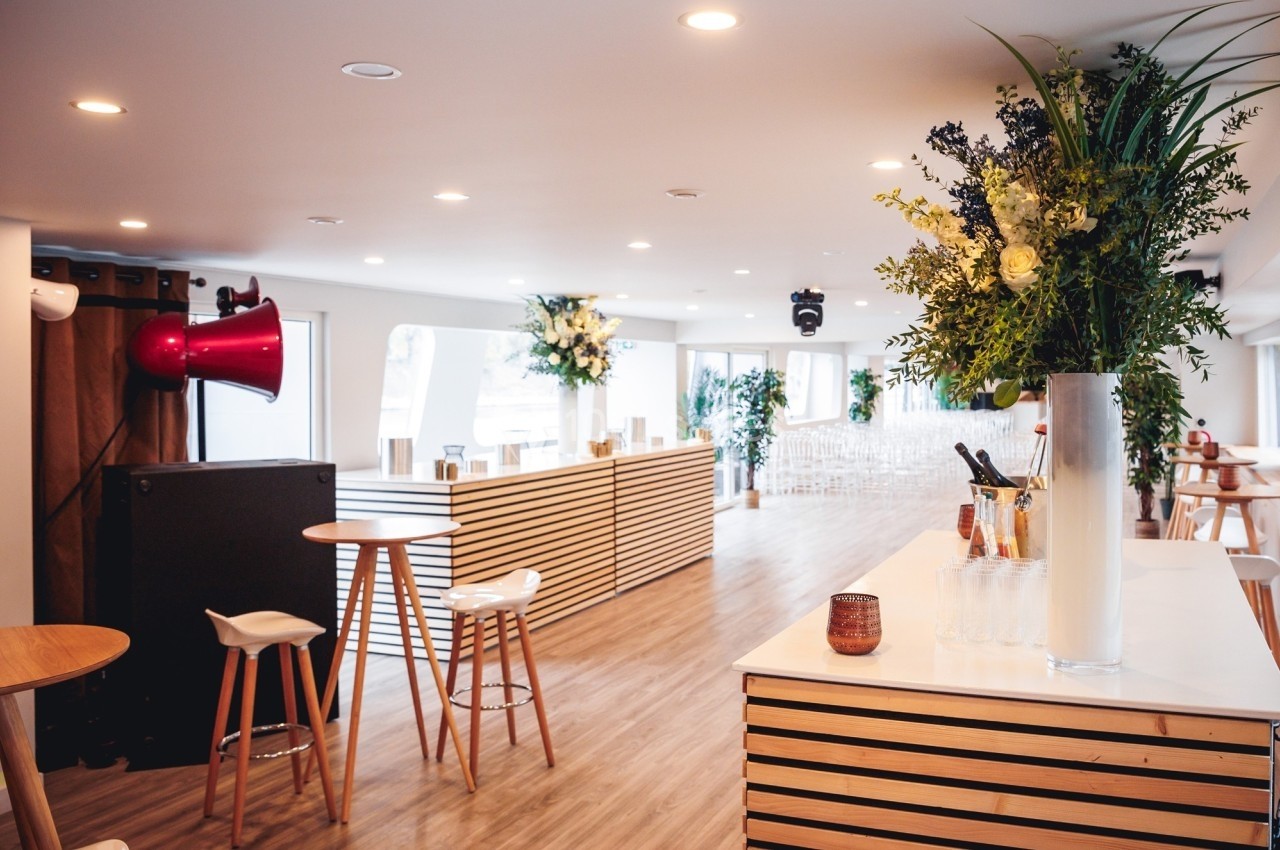 Salle lumineuse avec comptoirs en bois, tabourets, plantes décoratives et disposition pour un événement.