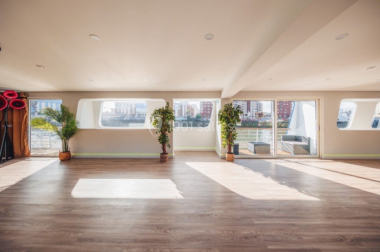 Salle lumineuse avec parquet, grandes fenêtres donnant sur un paysage urbain et des plantes en pot.