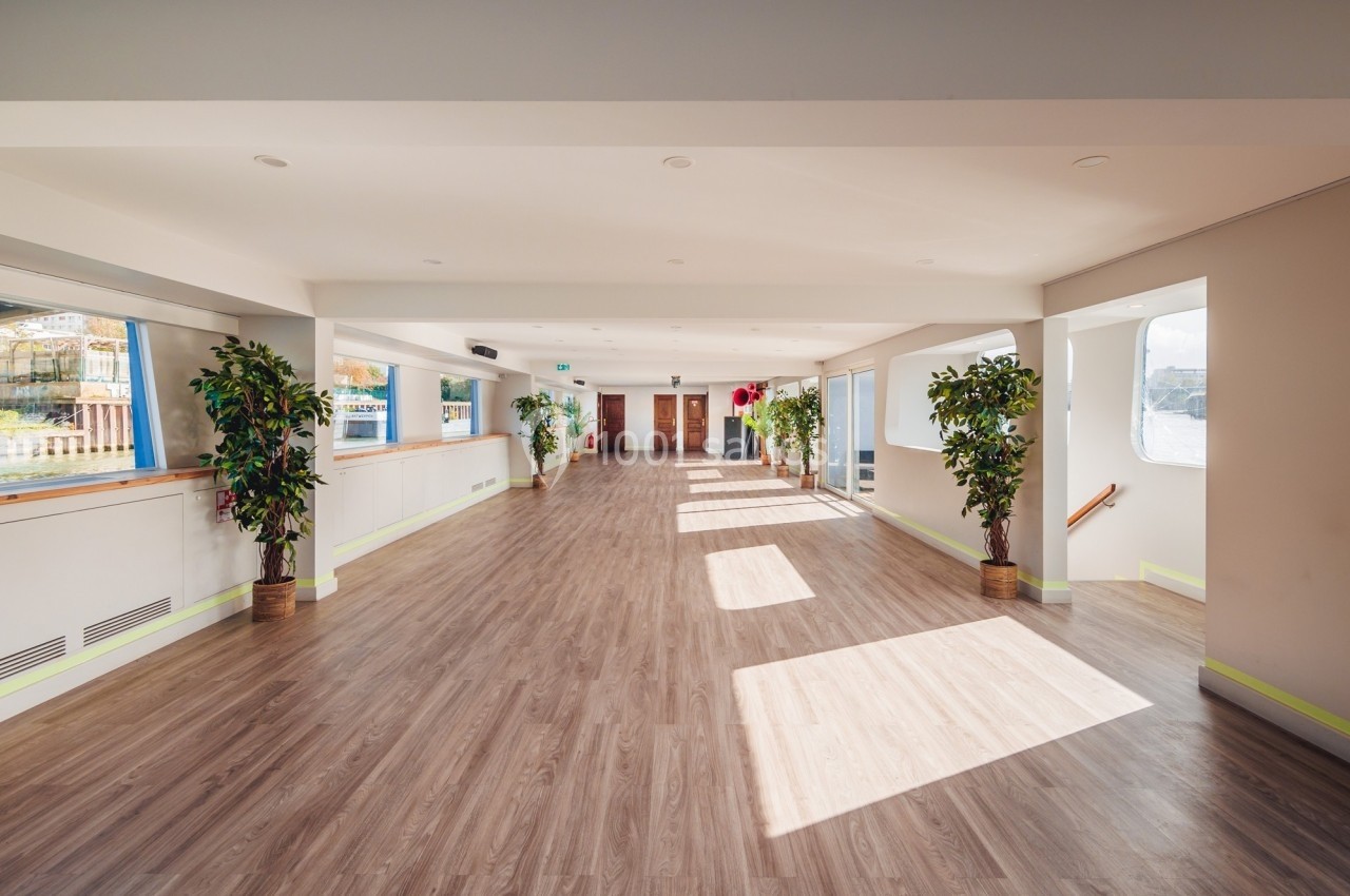 Salle lumineuse avec parquet, grandes fenêtres laissant entrer la lumière naturelle, et plantes en pots disposées le long…