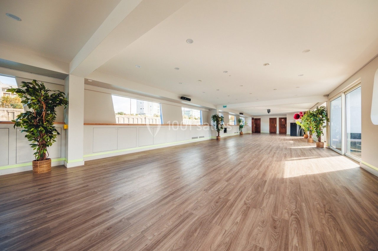 Salle spacieuse avec parquet en bois, grandes fenêtres laissant entrer la lumière et plantes décoratives.