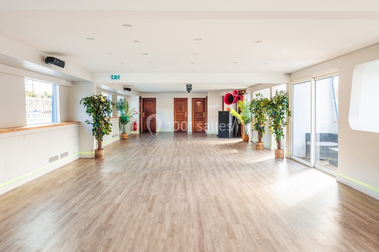Salle lumineuse avec parquet, plantes en pot, grandes fenêtres et portes en bois au fond.
