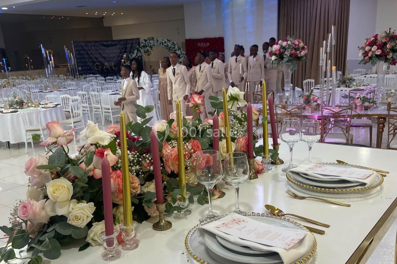 Salle de réception décorée avec des fleurs, bougies colorées et tables dressées, avec un groupe de personnes en arrière-plan.