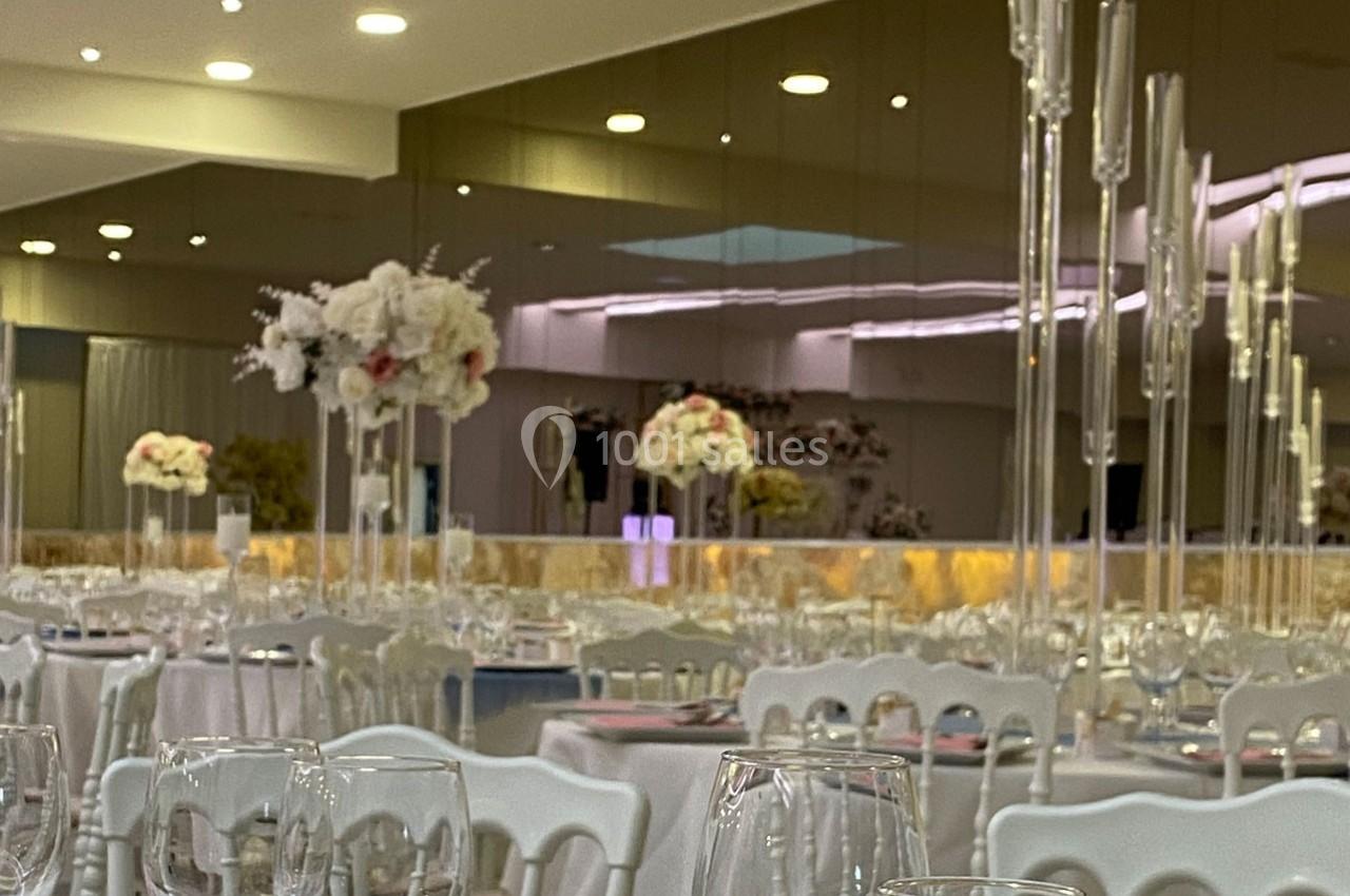 Salle de réception élégante avec tables dressées, décor floral blanc et rose, et chandeliers en verre.