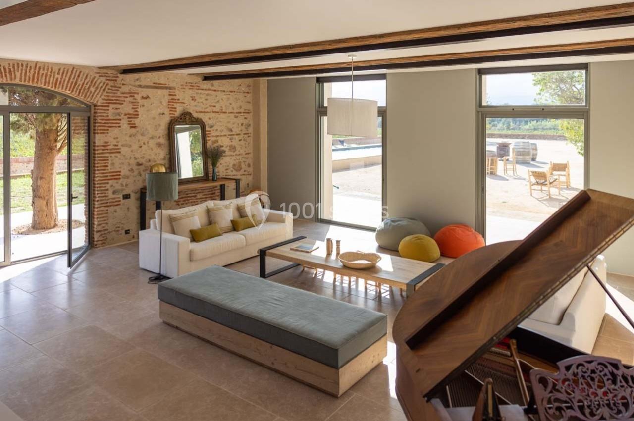 Salon lumineux avec murs en pierre, canapé, piano, poufs colorés et vue sur une terrasse extérieure.