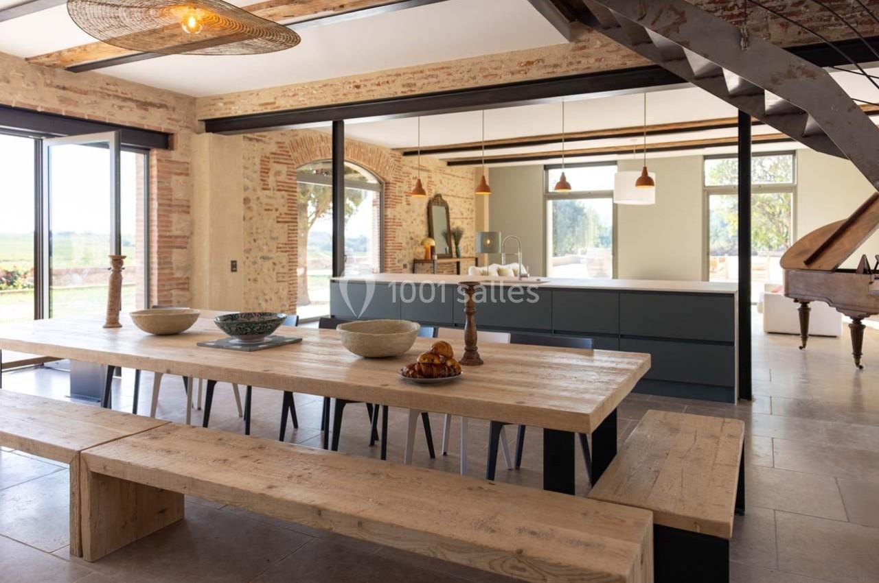 Salle à manger lumineuse avec une grande table en bois, cuisine moderne en arrière-plan et vue sur l'extérieur.