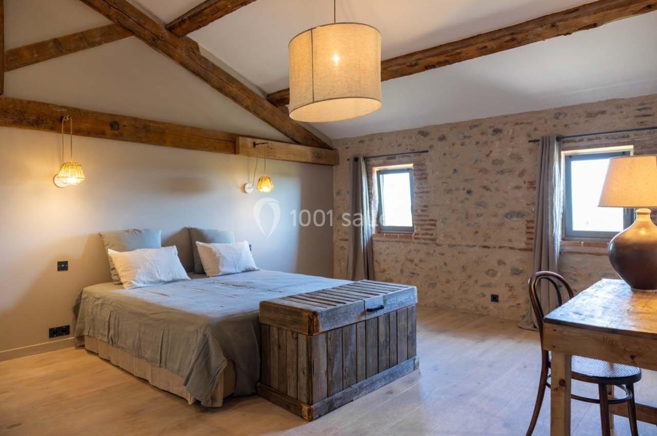 Chambre lumineuse avec poutres apparentes, lit double, coffre en bois, bureau et murs en pierre.