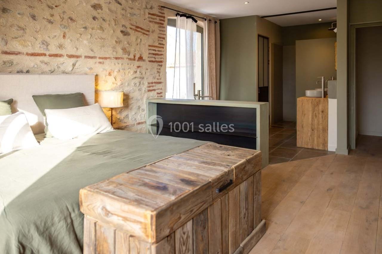 Chambre lumineuse avec mur en pierre, lit double, coffre en bois et salle de bain attenante avec lavabo moderne.