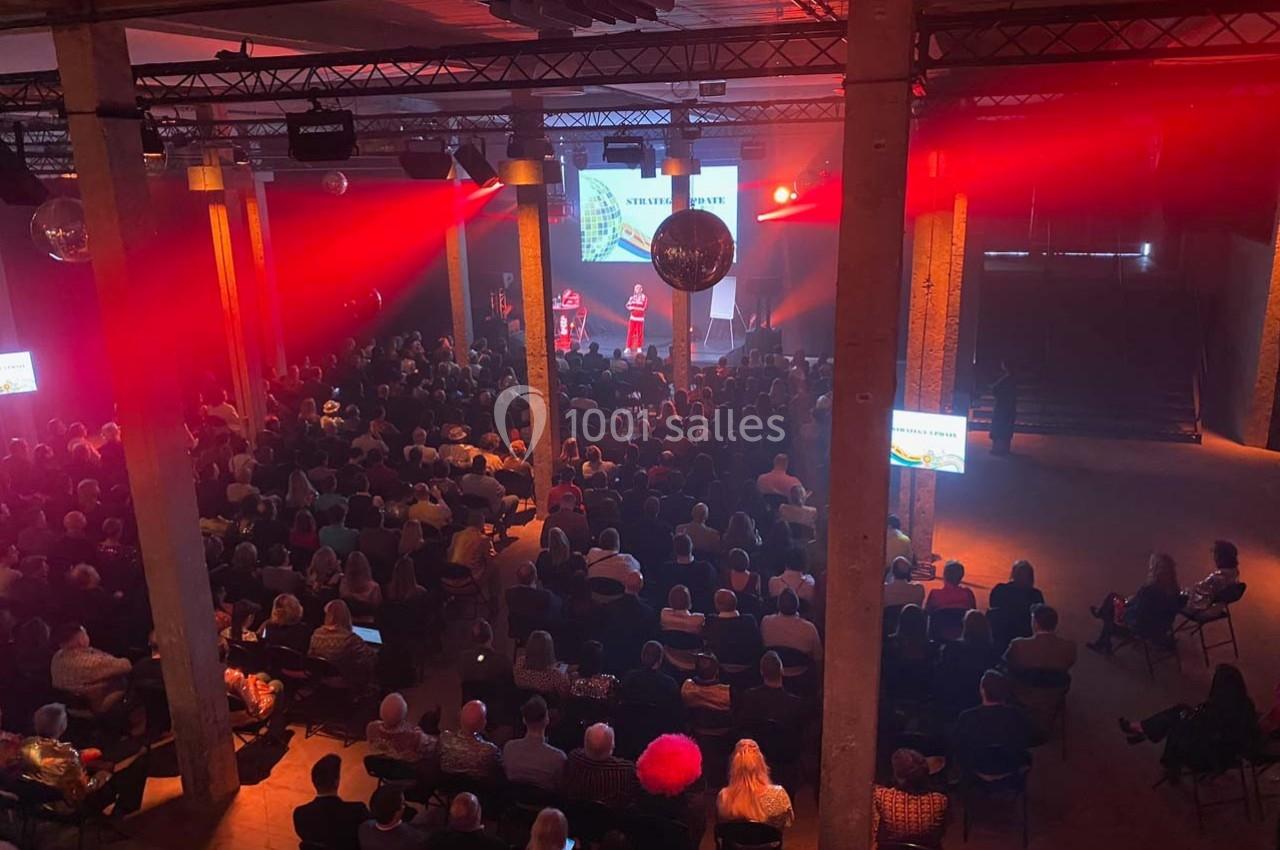 Salle sombre avec un public assis, éclairages rouges et scène illuminée où deux personnes présentent devant un écran.