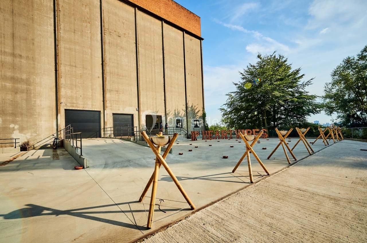 Cour extérieure avec sol en béton, structure en bois en premier plan et grand bâtiment industriel en arrière-plan.