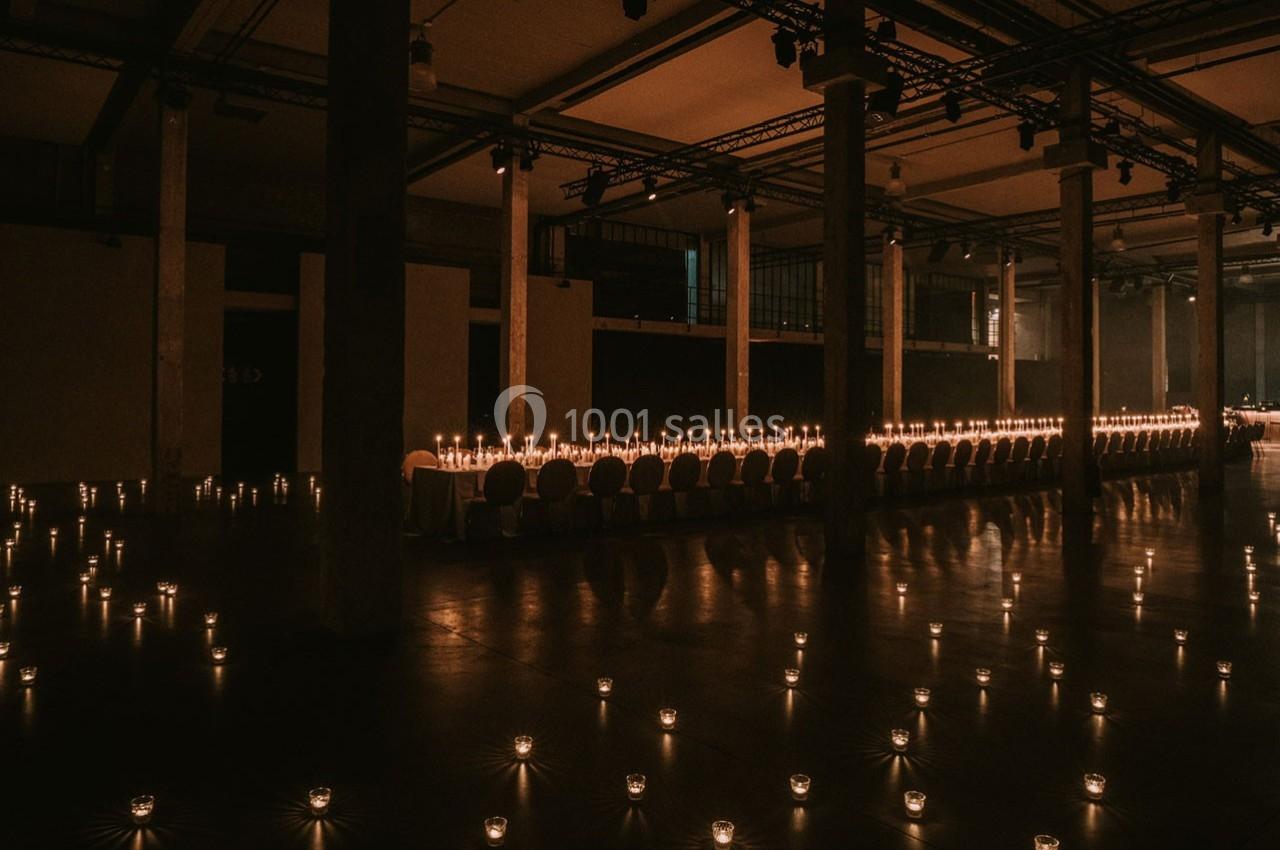 Grande salle sombre éclairée par des rangées de bougies sur une longue table et au sol, créant une ambiance chaleureuse.