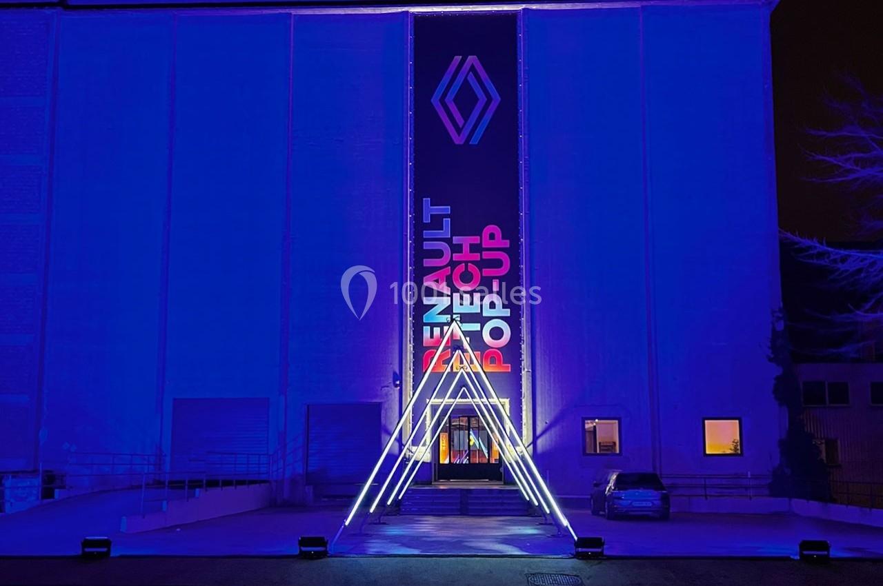 Façade d'un bâtiment illuminé en bleu avec un logo et une bannière ’Renault Tech Pop-Up’ au centre.