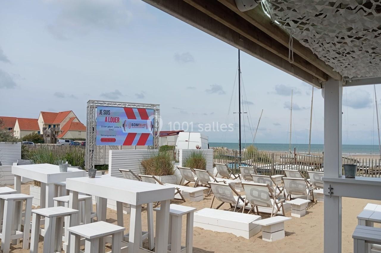 Terrasse de plage avec tables et chaises blanches, écran publicitaire, vue sur la mer et maisons en arrière-plan.