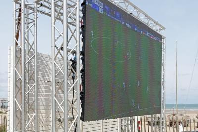 Écran géant installé en extérieur diffusant un match de football, avec une plage visible en arrière-plan.