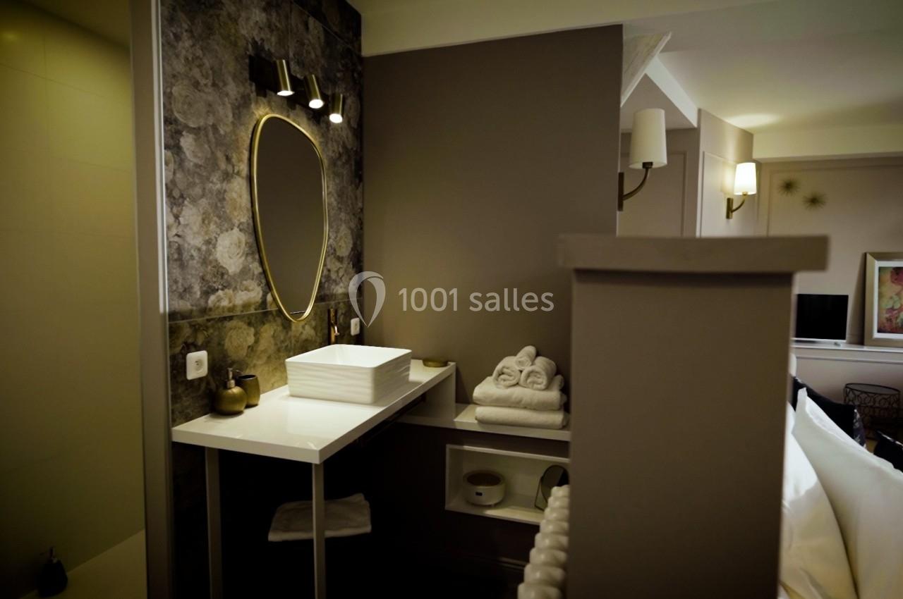 Salle de bain moderne avec lavabo blanc, miroir ovale doré, éclairage mural et serviettes pliées sur une étagère.