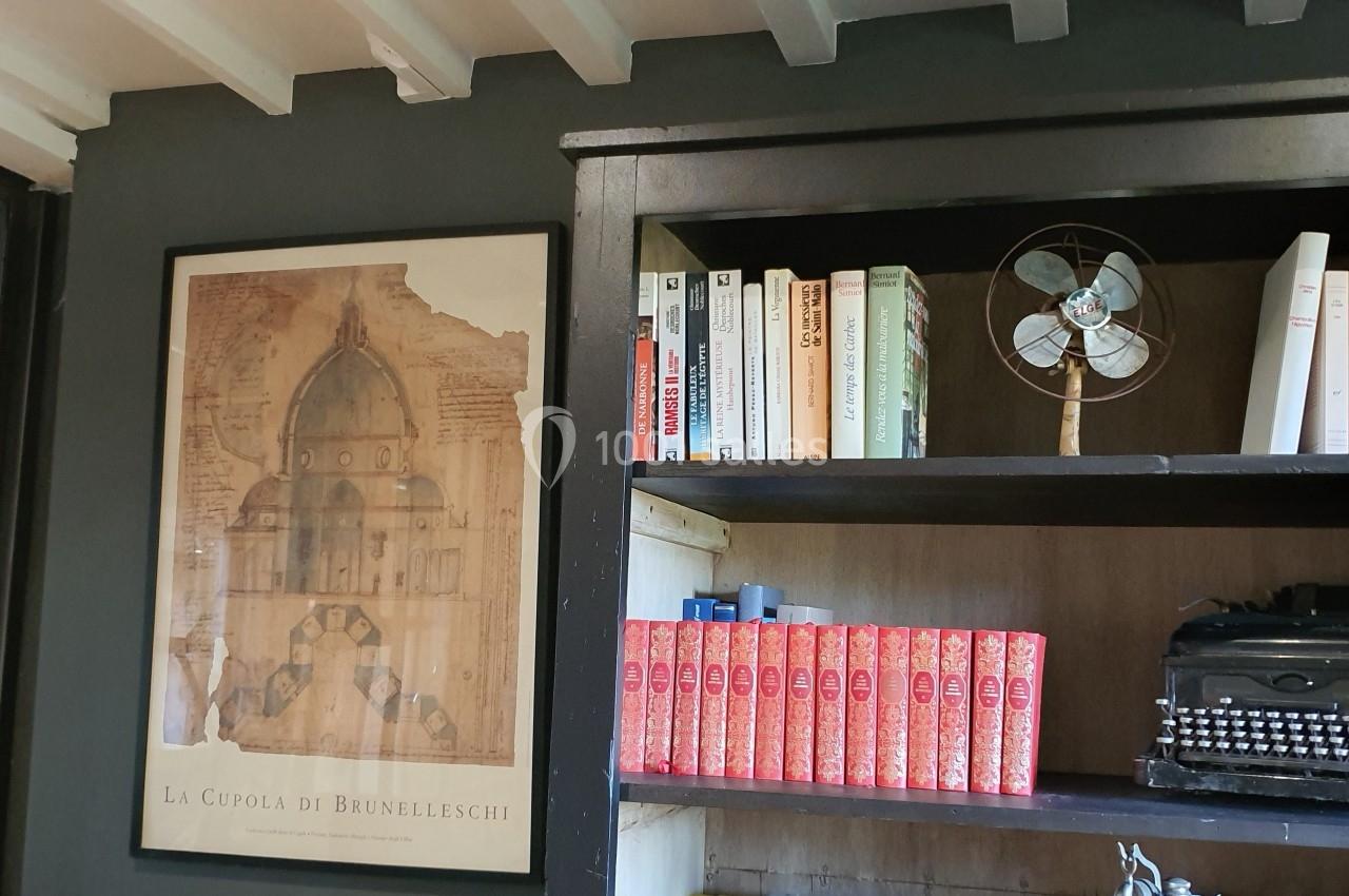 Affiche architecturale encadrée, étagère avec livres, ventilateur vintage et machine à écrire sur fond de mur sombre.