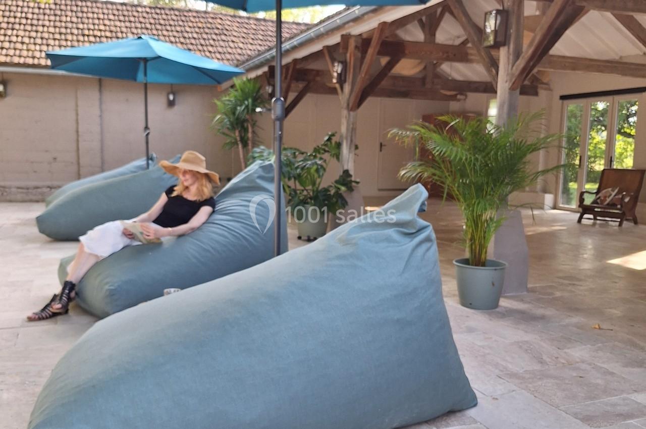 Femme assise sur un grand pouf bleu sous un parasol, sur une terrasse en pierre entourée de plantes.