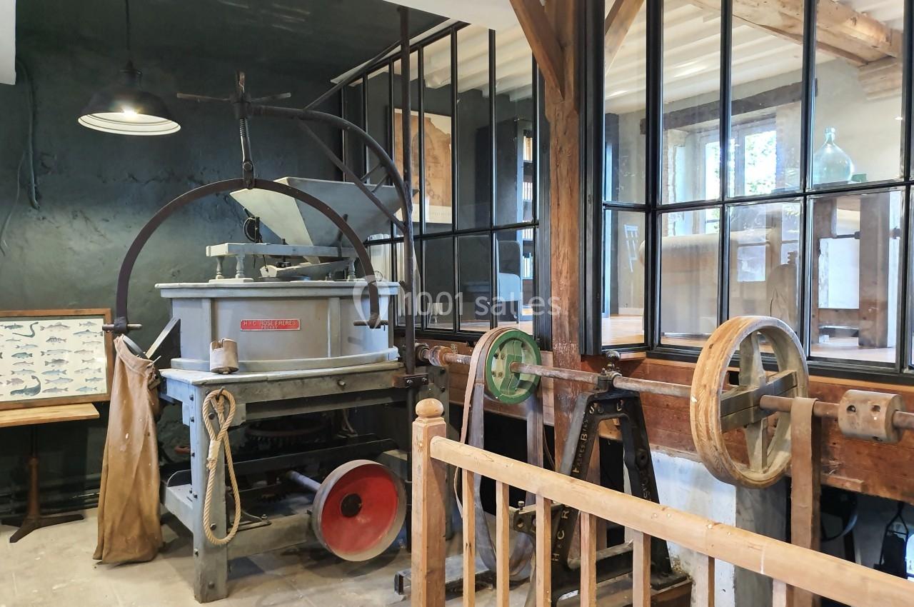 Machine ancienne dans un atelier avec poutres en bois, vitres séparant les espaces et éclairage suspendu.