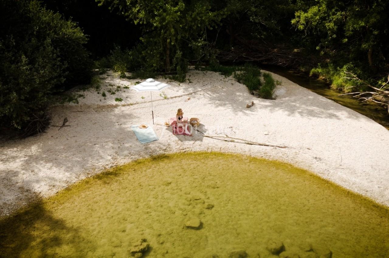 Une femme assise sur une plage de galets près d'une rivière, entourée de verdure et d'un parasol blanc.