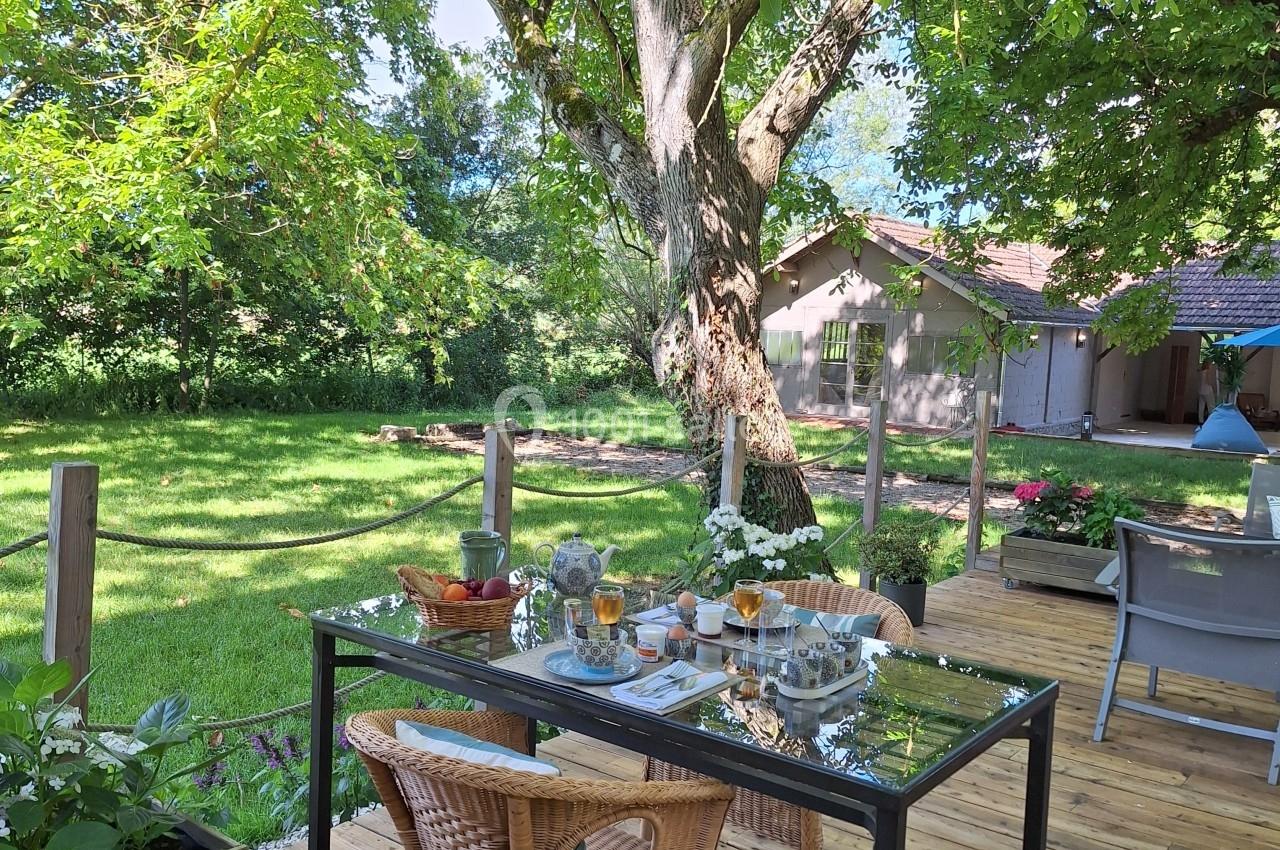 Table en verre dressée sur une terrasse en bois, entourée de chaises, avec vue sur un jardin verdoyant et arboré.