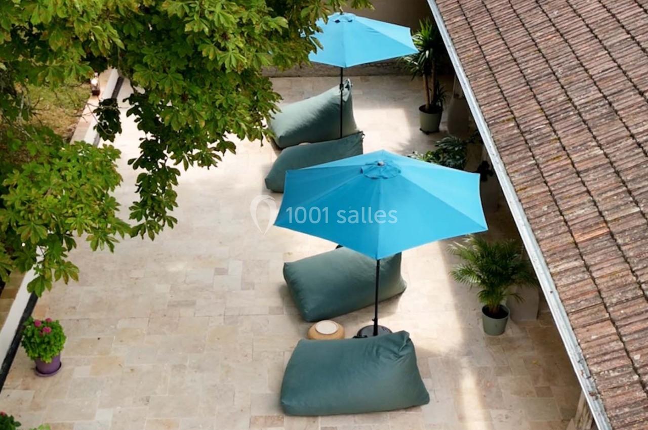 Terrasse en pierre avec parasols bleus, poufs verts et plantes en pot, entourée de végétation et d'un toit en tuiles.