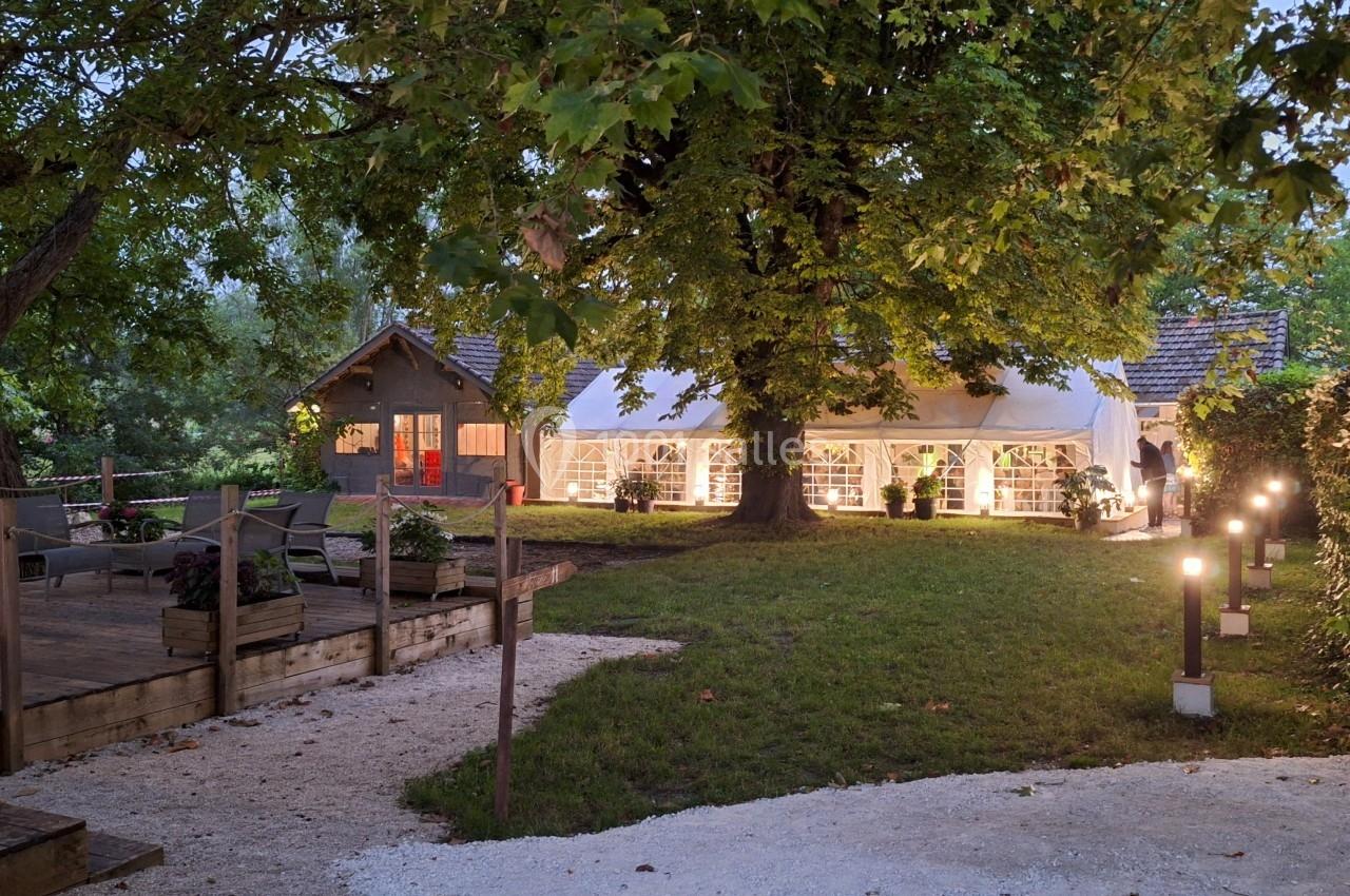 Jardin éclairé en soirée avec une grande tente blanche et une maison en arrière-plan sous des arbres feuillus.