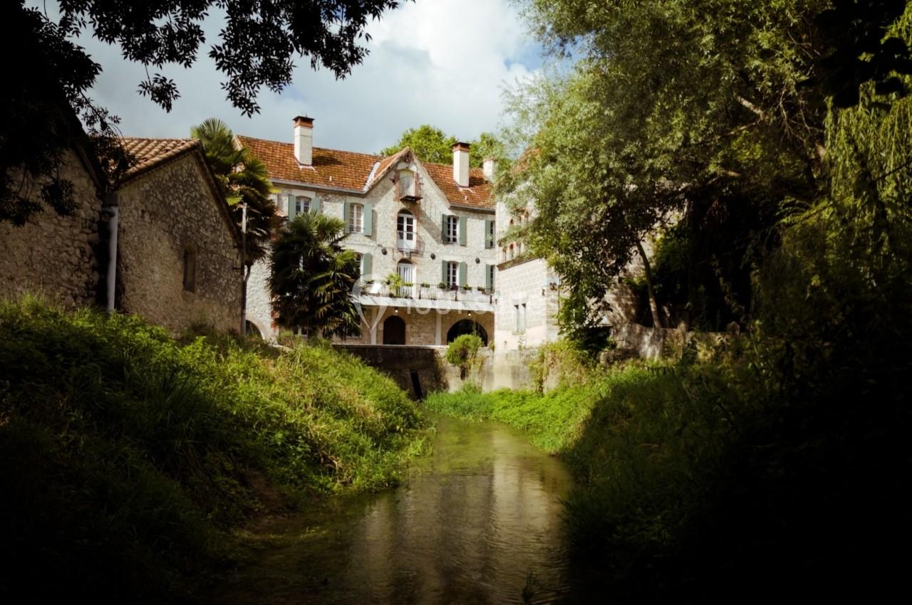 Grande maison en pierre avec toit en tuiles rouges, bordée d'arbres, surplombant un ruisseau entouré de verdure.