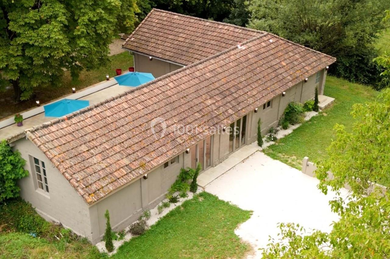 Bâtiment rectangulaire avec toit en tuiles, entouré de verdure, terrasse avec parasols bleus visible sur le côté.