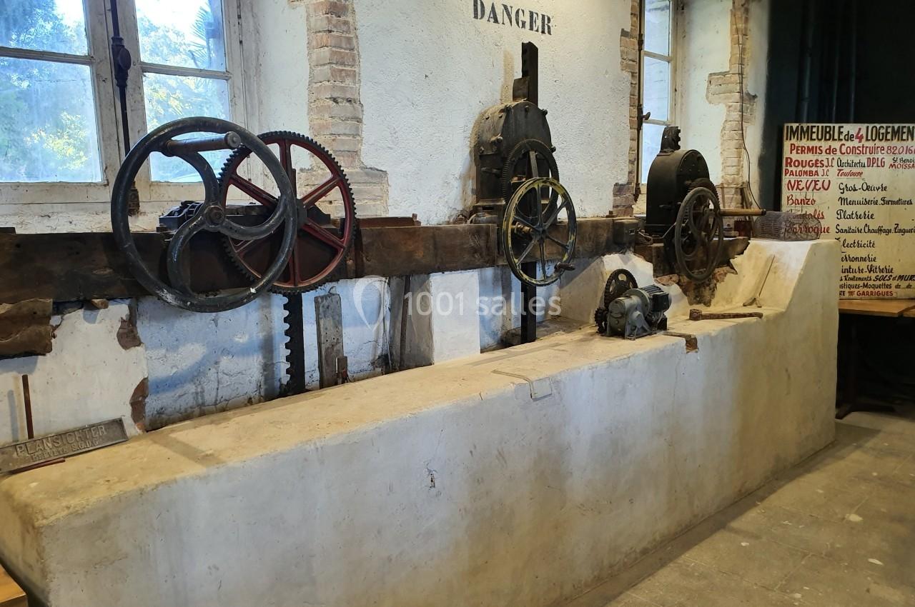 Ancien atelier avec mécanismes de courroies et engrenages fixés à un mur en pierre, éclairé par des fenêtres.