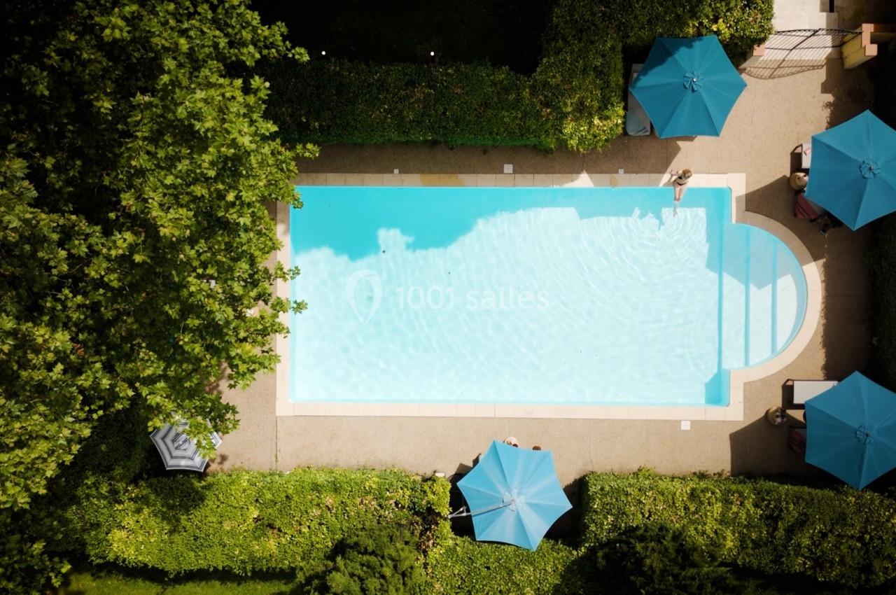 Vue aérienne d'une piscine entourée de verdure et de parasols bleus, avec une personne au bord de l'eau.