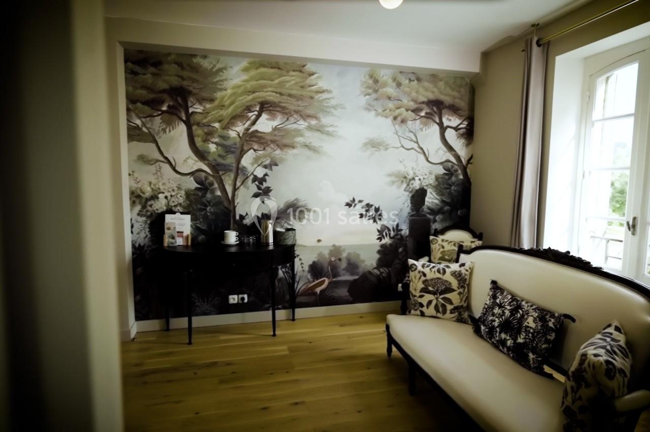 Salon avec canapé clair, table noire et mur décoré d'une fresque représentant un paysage naturel, éclairé par une fenêtre.