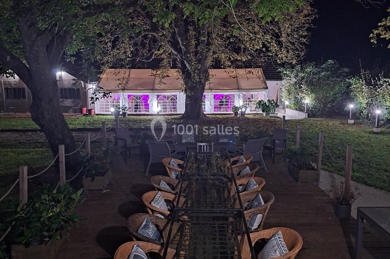 Terrasse en bois avec une longue table entourée de chaises, éclairée par des lumières, face à une tente illuminée la nuit.