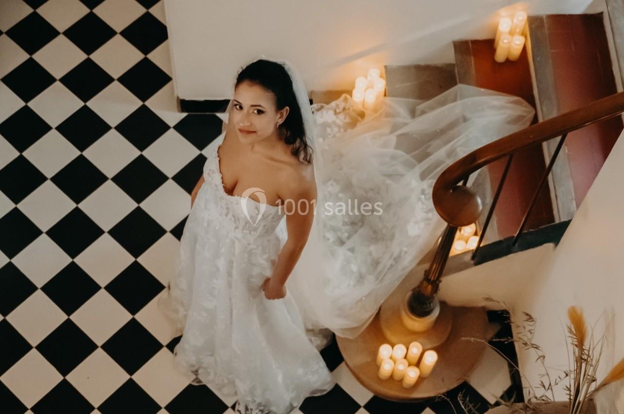 Femme en robe de mariée blanche debout sur un sol à carreaux noirs et blancs, entourée de bougies allumées.