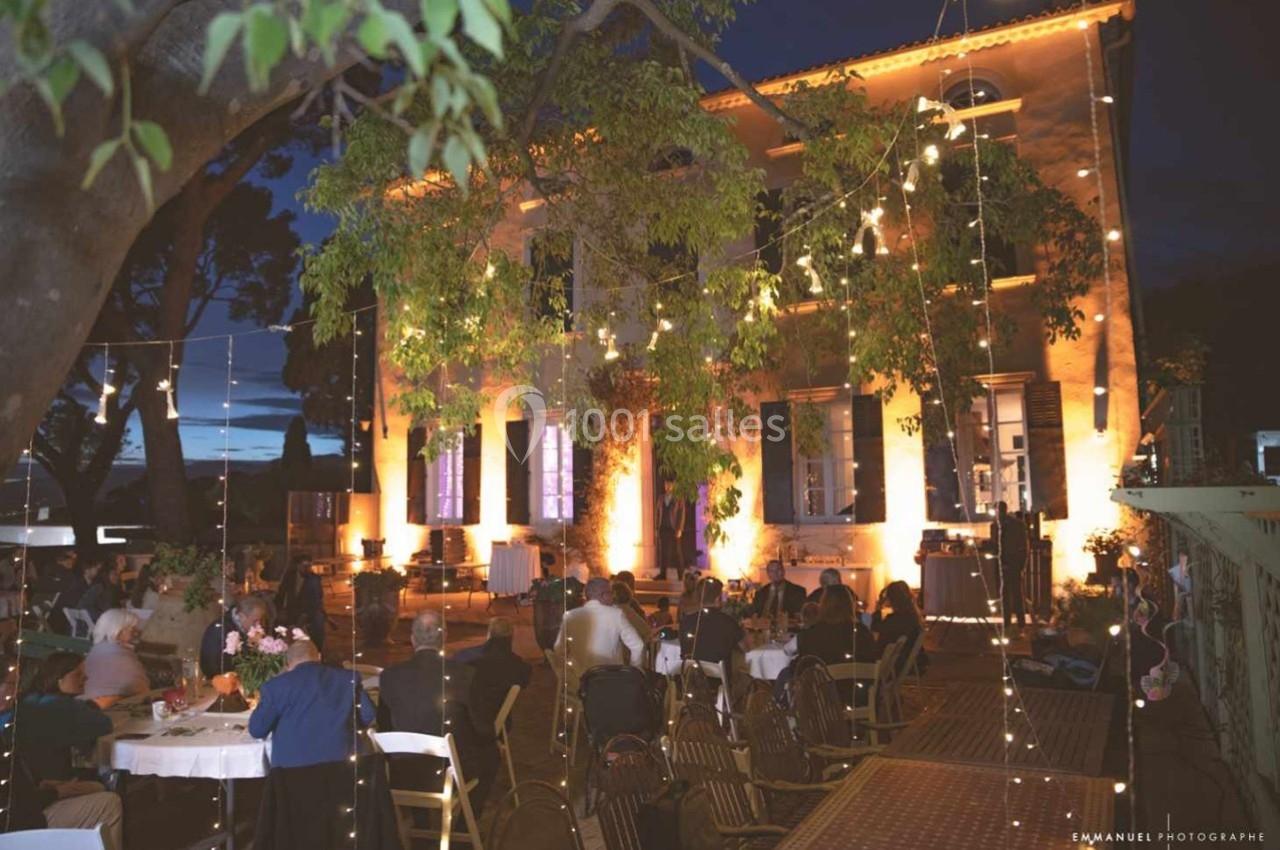 Façade d'une maison éclairée le soir, avec des guirlandes lumineuses et des invités assis à des tables en extérieur.