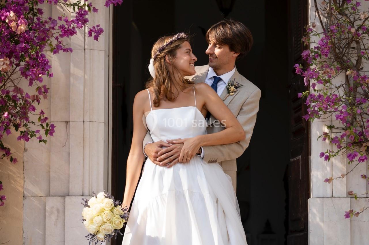 Un couple en tenue de mariage pose devant une entrée ornée de fleurs violettes.