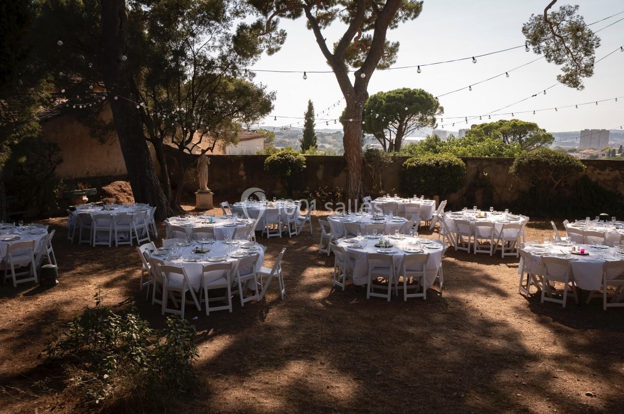 Tables rondes dressées avec nappes blanches dans un jardin ombragé, décoré de guirlandes lumineuses suspendues.